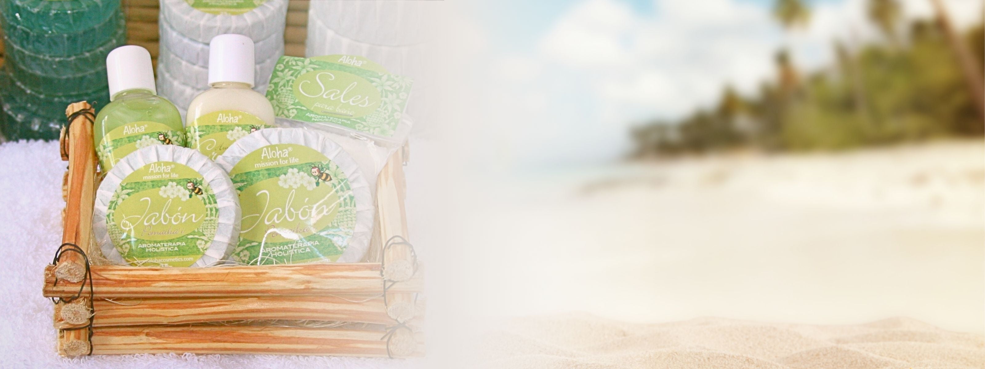 A beautifully arranged wooden gift basket filled with natural bath and body products sits atop a soft white towel. Wrapped herbal soaps, aromatic bath salts, and refreshing lotions from the Aloha mission for life collection are elegantly displayed, each adorned with green botanical packaging featuring a cheerful bee illustration. The scene transitions into a softly blurred tropical beach in the background, evoking a sense of relaxation, purity, and holistic well-being.