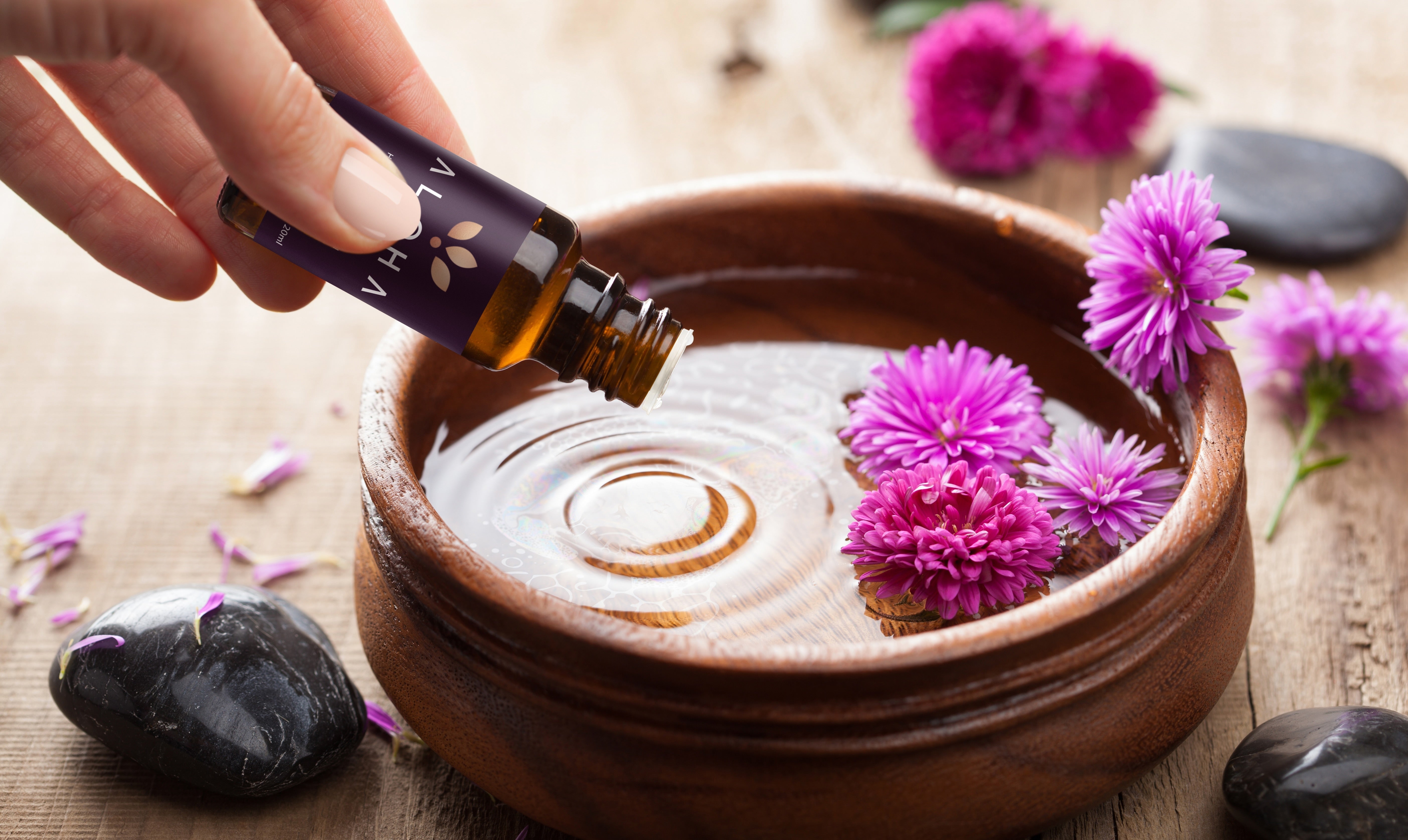 A hand pouring essential oil into a wooden bowl of water, surrounded by vibrant purple flowers, black stones, and scattered petals on a wooden surface